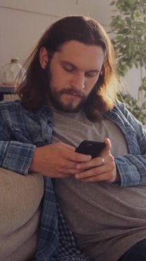 Ciddi bir adam evdeki sosyal medyada internetteki rahatlık ve mesajlaşma için telefon kullanıyor, rahat bir ev ortamında odaklanmış iletişimle meşgul oluyor. Dikey video.