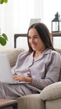 Mutlu bir kadın evde online eğlence ve eğlence için dizüstü bilgisayar kullanıyor, rahat ve rahat bir ortamda sanal alemde neşeli davranışlar sergiliyor. Dikey video.