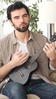 Ukulele çalan ve evde boş zamanlarında şarkı söyleyen, müzik eğitimi veren yaratıcı ve rahatlatıcı bir hobisi olan bir adam. Dikey video.