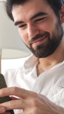 Evde çevrimiçi boş zamanlarında internet eğlencesiyle meşgul olan gülümseyen bir adam, rahat bir ortamda dijital aktivitelerden keyif alıyor. Dikey video.