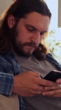 Cep telefonunu internette gezinmek ve internette sohbet etmek için kullanan düşünceli bir adam, rahat ve tanıdık bir yerde eğleniyor. Dikey video.