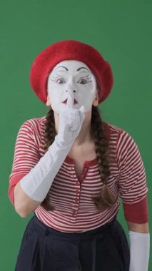 Komik kadın pandomimci yeşil arka planda sessiz ve etkileyici bir performansla sessizliği ve mahremiyeti işaret ediyor. Dikey video.