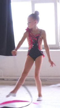 Stüdyoda bir kurdeleyle dinamik bir dans yapan kız, çağdaş koreografiyi sergiliyor ve sanatsal bir adanmışlıkla canlı bir performans sergiliyor. Dikey video.