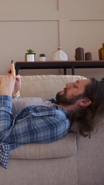 Evde dijital eğlence için telefon kullanan bir adam, rahat bir ortamda dijital aktiviteleri gevşeterek eğlence ararken can sıkıntısı çekiyor. Dikey video.
