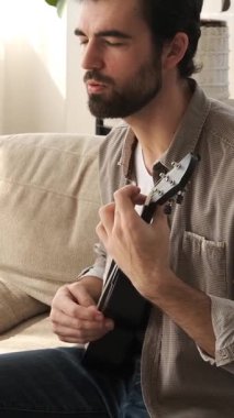 Evde öğrenirken gitar çalıp şarkı söyleyen, rahatlama ve memnuniyeti kucaklayan, rahat bir ortamda huzurlu bir müzik deneyimi yaratan içerikli bir adam. Dikey video.