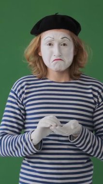 Üzgün pandomimci ağlayarak ve yeşil arka planda gözyaşlarını tutarak, hayal kırıklığını ve duygusal derinlik sorununu dışa vuruyor. Dikey video.