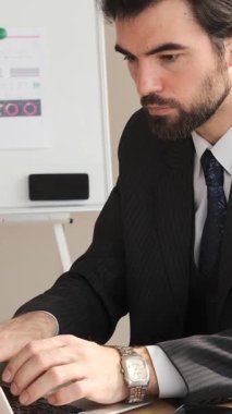 Ofiste dizüstü bilgisayarla çalışan konsantre işadamı, açıklamaları geliştiriyor ve analiz ediyor, profesyonel bir çalışma alanındaki stratejik görevlere odaklanıyor. Dikey video.