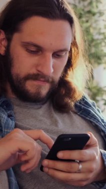 Memnuniyet duyulan bir adamın portresi cep telefonuyla internette geziniyor ve evde sohbet ediyor, rahatlamış ve içerik davranışlarıyla dijital iletişim kuruyor. Dikey video.