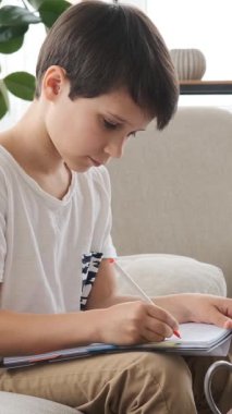 Düşünceli çocuk evde bir defterde ödevlerini yapıyor, odaklanmış ve sessiz bir ortamda e-öğrenme yoluyla eğitime hazırlanıyor. Dikey video.