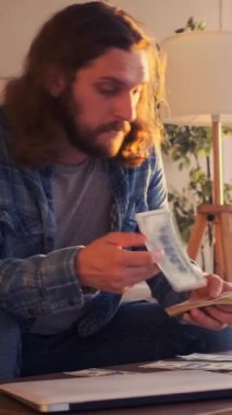 Düşünceli bir adam evde nakit para sayıyor, refah, gelir ve krediyi düşünüyor, sakin bir iç çevrede finansmanı düşünceli bir şekilde yönetiyor. Dikey video.