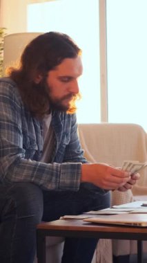 Kafası karışmış bir adam evde nakit para ya da kâr sayıyor, sermaye, maaş ve bütçe yönetiyor, özel bir iç alandaki mali sonuçları yansıtıyor. Dikey video.