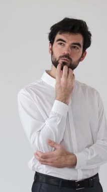 Düşünceli ve şüpheci bir adam beyaz bir arkaplanda beyin fırtınası yapar ve düşünür. Hayal gücü ve düşünceli bir ifadeyle bir soruyu yansıtır. Dikey video.