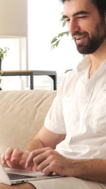 Bilgisayarla online çalışan mutlu bir adam, evde video görüşmesi yapan, sohbet ve bağlantıyla ilgilenen, rahat bir ortamda iletişimi teşvik eden. Dikey video.