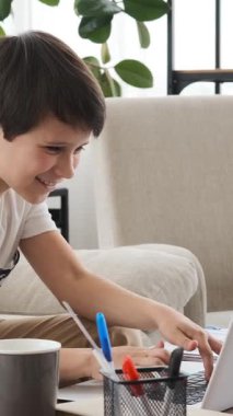 Dost canlısı bir çocuk ev ödevi olarak dizüstü bilgisayar kullanıyor ve evde gülüyor, rahat ve neşeli bir çalışma ortamında bir öğrenci olarak dersten zevk alıyor. Dikey video.