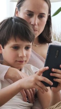 Dost canlısı bir oğul ve anne internet eğlencesi ve evde online eğlence için telefon kullanıyor, sıcak bir ortamda dijital zevki paylaşıyorlar. Dikey video.