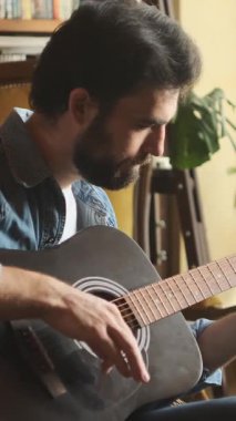 Evde gitar çalan mutlu bir adam, eğlence ve eğlenceyle meşgul, rahat ve evcimen bir ortamda eğlenerek bir performans sergiliyor. Dikey video.