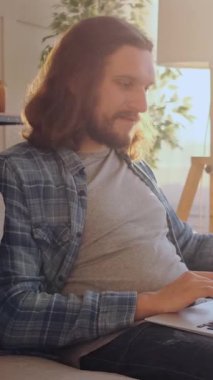 Neşeli bir adam, evde akşamları bira içerken internetten bilgisayarla sohbet ediyor, canlı ve bağlı dijital bir varlıkla eğleniyor. Dikey video.