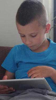 Gülümseyen çocuk çevrimiçi eğlenceye dalmış, evde bir tablette video oyunları oynuyor, canlı ve dijital bağlantının tadını çıkarıyor. Dikey video.