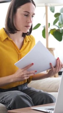 Evinde dokümanlar ve dizüstü bilgisayarlarla çalışan düşünceli serbest çalışan, odaklanma ve özveriyle uzak bir işle meşgul olan biri. Dikey video.