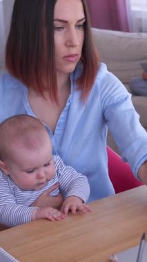 Dost canlısı bir kadın evde dizüstü bilgisayarla çalışıyor, kucağında bir bebek tutuyor, ev ortamında sıcak bir şekilde iş bulma görevlerini yönetiyor. Dikey video.