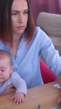 Ciddi bir kadın evde bir bebeği tutarken internetten çalışıyor, konsantrasyon ve bağlantıyı yönetiyor, ev ortamında denetleme görevleri yapıyor. Dikey video.