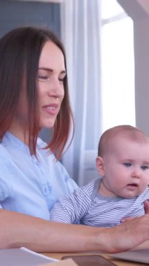 İyimser bir anne evde çalışır, kucağında bir bebek, iletişim ve muhasebe işlerini coşkuyla yürütür. Dikey video.