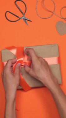 Kadın hediye paketi hazırlıyor ve turuncu arka planda bir kurdele bağlıyor. Sürpriz bir kutlamaya özenle ve dikkatle bir hediye hazırlıyor. Dikey video.
