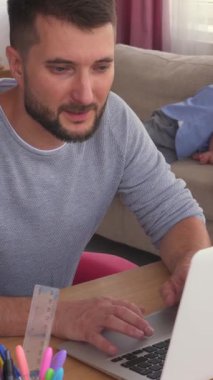 Çocuklar arka planda oynarken evde dizüstü bilgisayar kullanarak çalışan, aile ve ev hayatını rahat ve hayat dolu bir ortamda dengeleyen bir adam. Dikey video.
