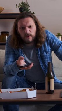 Bir portrede mutlu bir adam televizyon izlerken gülüyor, pizza yiyor ve akşam eğlencesinde bira içiyor, neşe içinde dinleniyor. Dikey video.