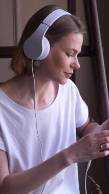 Hafta sonu evde boş zamanlarında telefon kullanan ve müzik dinleyen iyimser bir kadın, neşeli bir atmosferde eğleniyor. Dikey video.