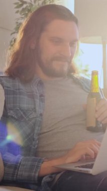 Keyifli bir adam akşam evde online eğlence için dizüstü bilgisayar kullanıyor, rahat bir atmosferde dijital eğlenceye dalarken birasını yudumluyor. Dikey video.