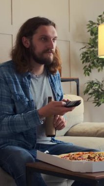 Mutlu bir adam televizyon izliyor, pizza yiyor ve akşamları evde bira içiyor. Neşeli ve rahat bir ortamda dinleniyor. Dikey video.