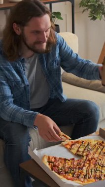 Akşam evde televizyonla eğlenirken bira içen ve pizza yiyen olumlu bir adam neşeli ve rahat bir ev ortamında gevşiyor. Dikey video.