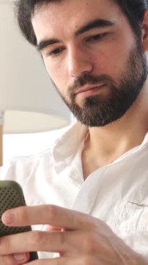 Evde dinlenirken çevrimiçi boş zaman ve mesajlaşma için cep telefonu kullanan, neşeli bir şekilde dijital iletişim kuran olumlu bir adam. Dikey video.