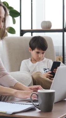Düşünceli bir anne evde dizüstü bilgisayarla çalışırken, oğlu da yakınlarda bir telefon ile vakit geçirip iş ve aile zamanını dengeliyor. Dikey video.