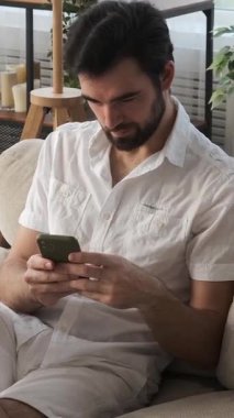 Mutlu bir adam evde sosyal medyada sohbet etmek için cep telefonu kullanıyor, iletişimi ve rahatlamayı kucaklıyor, rahat bir ortamda dijital olarak bağlanıyor. Dikey video.