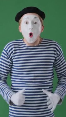 Şok olmuş pandomimci adam yeşil bir arka planda başparmağını gösterip alkışlıyor, anlamlı kutlama jestleriyle şoka tepki veriyor. Dikey video.