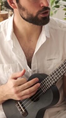 Gitar ve ukulele çalan heyecanlı bir adam, evde boş zamanlarında müzik söyleyen, memnuniyet saçan, canlı ve doyurucu bir müzik aktivitesi yapan. Dikey video.