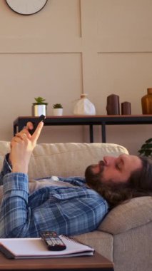 Evde cep telefonuyla eğlenirken sosyal medyada mesaj atan, dijital ortamda bağlantı ararken sıkıntıdan boğulan bir adam. Dikey video.