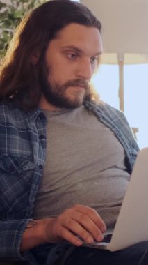 Akşamları evde dizüstü bilgisayarla çalışan özenli bir adam, rahat bir çalışma alanı içinde odaklanma ve adanmışlıkla iletişim kuruyor. Dikey video.