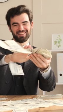 Ofiste çalışırken para sayıp saçan gülen işadamı, kazanç ve karı kutlayan, profesyonel bir çalışma alanında neşeli bir finansal an yaşıyor. Dikey video.