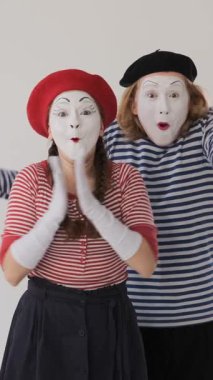 Erkek ve kadın pandomimciler şaşkınlık dolu ifadelerle alkışlıyor, beyaz bir arkaplanda hayret haykırıyorlar, dinamik jestlerle şaşkınlıklarını iletiyorlar. Dikey video.