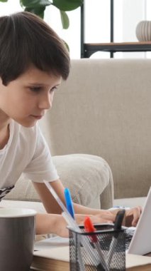 Mutlu çocuk evde ev ödevlerini öğrenmek ve düşünmek için dizüstü bilgisayar kullanıyor, rahat bir eğitim alanında hevesle çalışıyor. Dikey video.