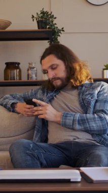 Sıkılmış bir adam cep telefonunu online eğlence için kullanıyor ve evde internette geziniyor, tanıdık bir ortamda monotonluğu hafifletmek için eğlence arıyor. Dikey video.