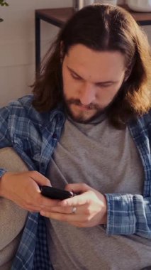 Evde boş zamanlarında akıllı telefon kullanan, rahat bir ortamda rahatlamaya ve dijital etkileşime odaklanan ciddi bir adam. Dikey video.