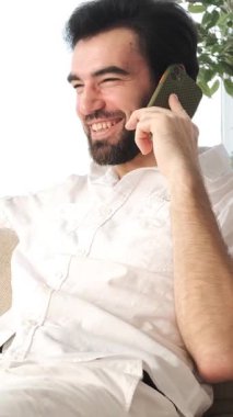 Memnuniyet verici bir adam evdeki bir konuşma sırasında cep telefonu kullanıyor, telekomünikasyon ve dedikodularla uğraşıyor, rahat bir ortamda iletişimi teşvik ediyor. Dikey video.