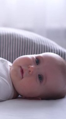 Masum bir bebek, evde yatakta yatarken saf mutlulukla gülüyor, emekleme çağında boş zamanların ve eğlencenin tadını çıkarıyor. Dikey video.