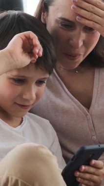 Neşeli anne ve oğul evde cep telefonuyla çevrimiçi boş zamanlarında eğleniyor ve neşeli bir atmosferde iletişimin tadını çıkarıyor. Dikey video.