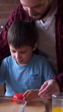 Bakıcı baba ve okul çocuğu evde sebze doğruyor ve salata hazırlıyor. Odaklanarak anlamlı bir aşçılık görevini paylaşıyorlar. Dikey video.