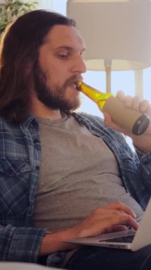 Arkadaş canlısı bir adam evde dizüstü bilgisayarla online eğlence yaparken bira yudumluyor sıcak ve rahat bir ortamda internet keyfi yapıyor. Dikey video.